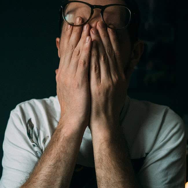 Close-up of a man indoors with hands covering his face, expressing emotions like sadness or disappointment.